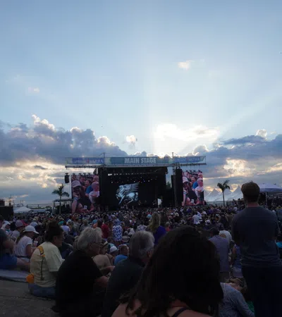 Huge crowd gathered at an outdoor music festival facing the main stage under a partly cloudy sky at sunset.