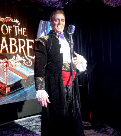 Performer in gothic costume stands next to a microphone with Tales of the Macabre sign behind him on stage.