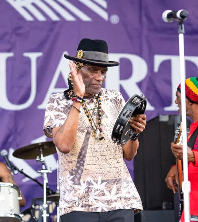 Musicians performing on stage with tambourine and drums, vibrant outfits, and purple backdrop.