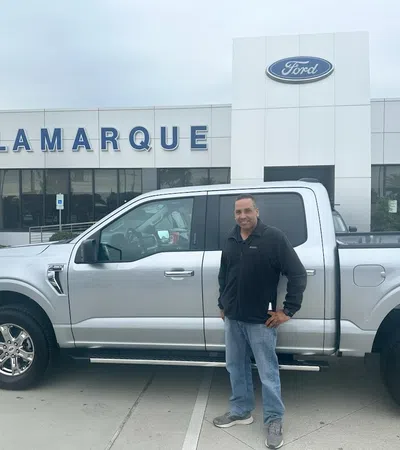 Man standing beside a silver Ford pickup truck in front of Lamarque Ford dealership on a cloudy day
