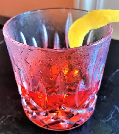 Close-up of a classic red cocktail in a textured glass with a lemon twist garnish on dark surface.