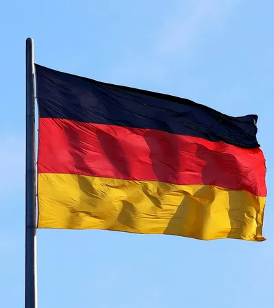 German flag with black, red, and yellow stripes waving on a flagpole against a clear blue sky.