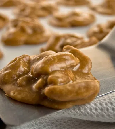 Close-up of freshly made praline candy clusters on a baking sheet being lifted with a metal spatula.