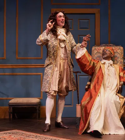 Three actors in 18th-century costumes performing on stage with blue paneled walls and period furniture.
