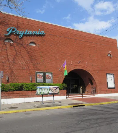Red brick building with arched entrance and Prytania sign under blue sky, surrounded by greenery and street elements.