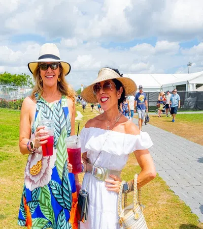 Two women in summer dresses and hats holding drinks at New Orleans Jazz Festival under bright sky.