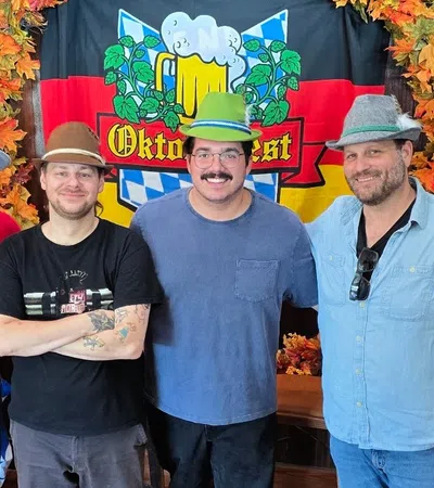 Four men wearing festive hats pose smiling in front of an Oktoberfest banner with autumn leaves decorations.