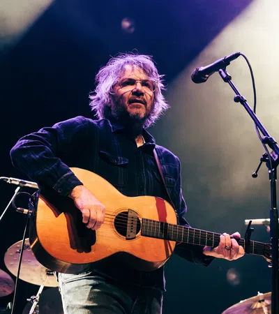 Musician playing acoustic guitar on stage with microphone and colorful stage lighting in smoky background.