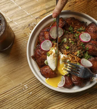 Bowl of fried plantains with poached egg, shredded meat, and radish slices on a wooden table with iced tea.