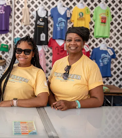 Three smiling women standing behind a counter with colorful t-shirts hanging on the wall behind them