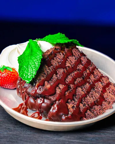 Decadent chocolate cake slice topped with chocolate ganache, whipped cream, fresh strawberry, and mint leaves on a white plate.