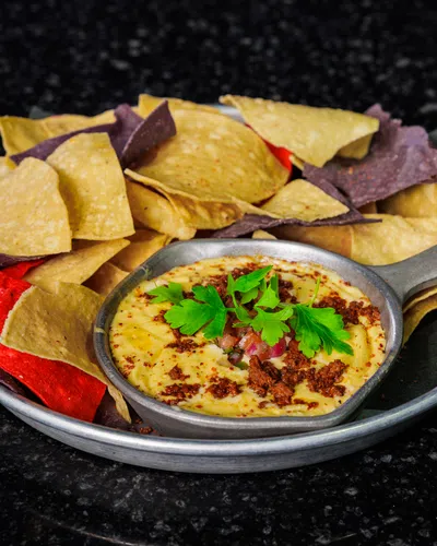 Metal plate with multicolored tortilla chips and creamy dip garnished with parsley on a black surface