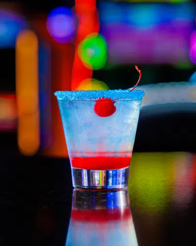 Cocktail glass with blue sugar rim, cherry garnish, and layered red drink on a reflective bar counter with blurred colorful lights.