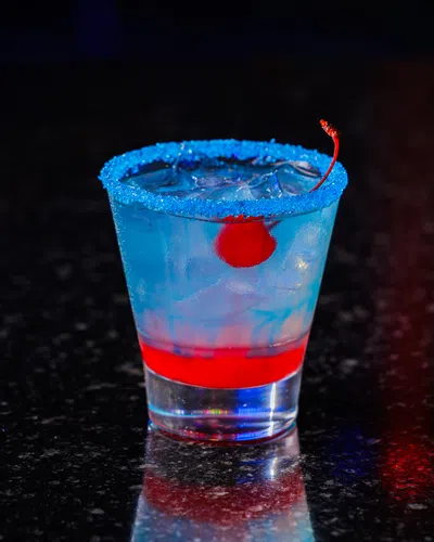 Blue cocktail with ice, red cherry, and red syrup at the bottom in a glass with blue sugared rim on dark surface.