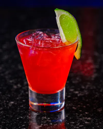 Red cocktail drink with ice cubes and a lime wedge garnish on a dark granite countertop.