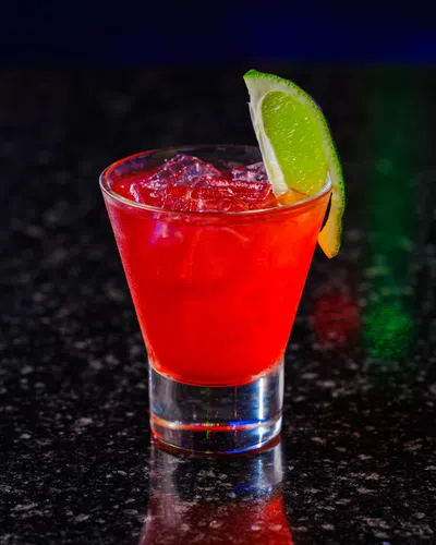 Red cocktail with ice and lime wedge on dark granite bar counter under low light.