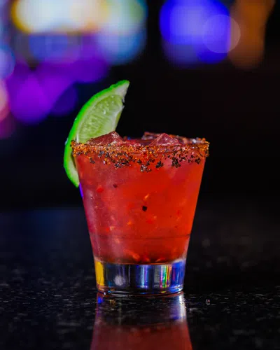 Spicy red cocktail with chili-rimmed glass and lime wedge on dark countertop with colorful bokeh background
