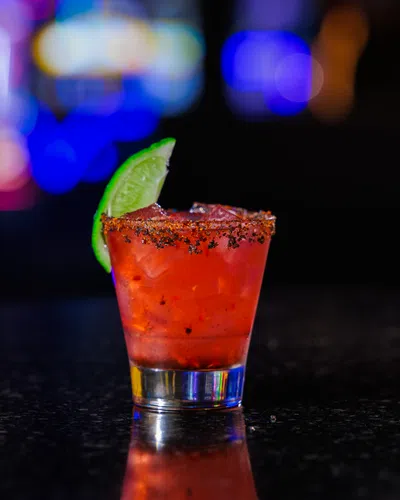 Red cocktail with ice and lime wedge on rim, garnished with chili powder, on black countertop with blurred lights.