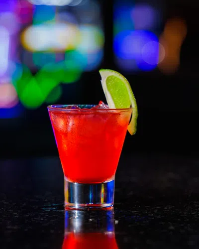 Bright red cocktail with ice and lime wedge on dark bar counter with colorful blurred lights
