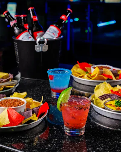 Plates of colorful tortilla chips with dips and cocktails on a dark bar table with bottles in a bucket in background