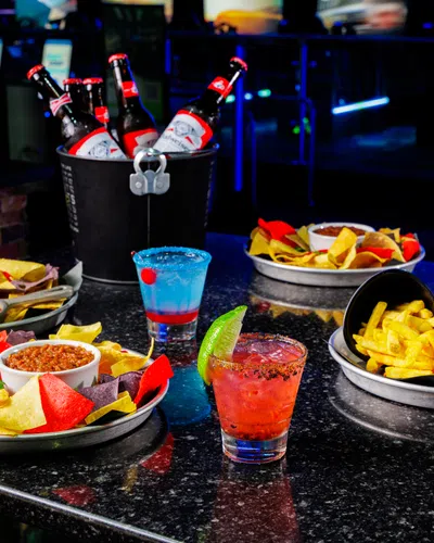 Plates of nachos, fries, sandwich, colorful cocktails, and bucket of beer bottles on a dark table in a bar setting.