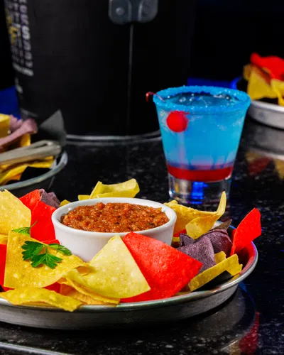 Colorful tortilla chips served with salsa and dip alongside vibrant cocktails on a dark table.