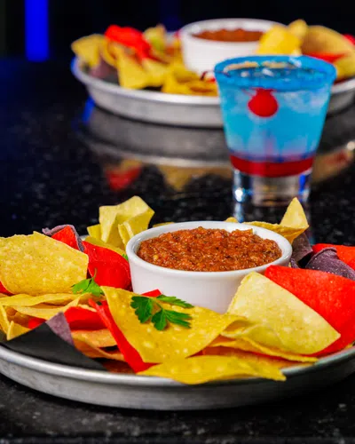 Plate of tri-color tortilla chips with salsa dip on a black table with colorful drinks in the background