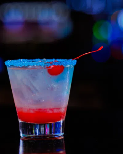 Cocktail glass with a blue sugar rim, ice, red syrup at the bottom, and a cherry garnish on a dark bar background.