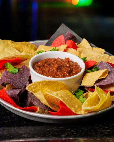 Plate of multicolored tortilla chips served with a bowl of chunky salsa on a dark table with colorful blurred background.
