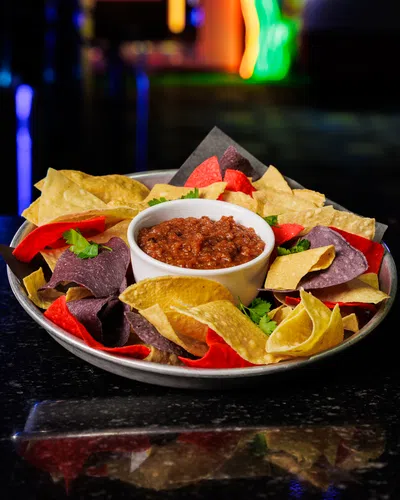 Plate of tri-color tortilla chips served with a bowl of chunky salsa on a dark reflective surface.
