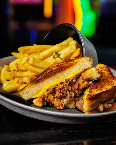 Grilled cheese sandwich with pulled meat and golden French fries served on a metal plate with blurred colorful background.