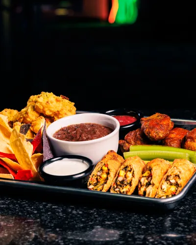 Tray with assorted appetizers including chips, dipping sauces, chicken wings, celery, and stuffed rolls on black surface.