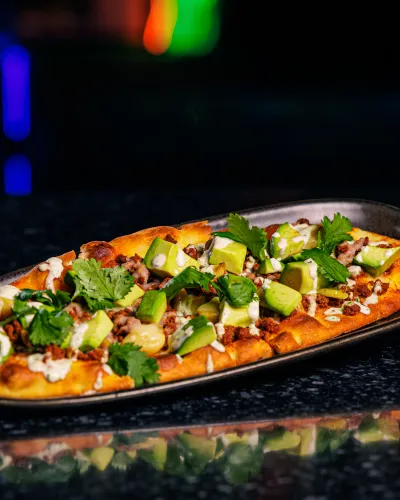 Flatbread topped with avocado, cilantro, ground meat, and sauce on a dark oval plate against a dark background