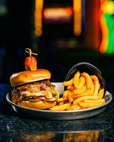 Juicy cheeseburger topped with a pepper and a side of golden curly fries on a metal tray with dark background.