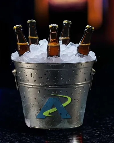 Metal bucket filled with ice and five brown beer bottles on a dark surface with blurred colorful background.