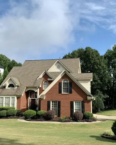 Brick and siding suburban house with manicured lawn, shrubs, and trees under a partly cloudy sky.
