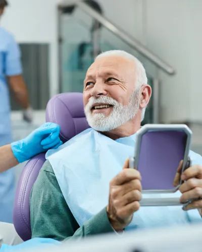 Smiling elderly man at dental clinic holding a mirror while dentist in mask and gloves talks to him