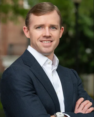 Professional man with light brown hair in dark suit and white shirt smiling outdoors with arms crossed.