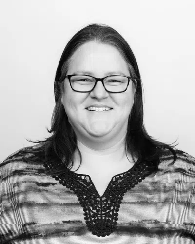 Smiling woman with glasses and patterned top posing against a plain light background in black and white photo