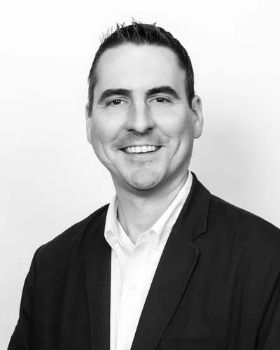 Black and white portrait of a smiling man in a white shirt and black blazer against a plain background.