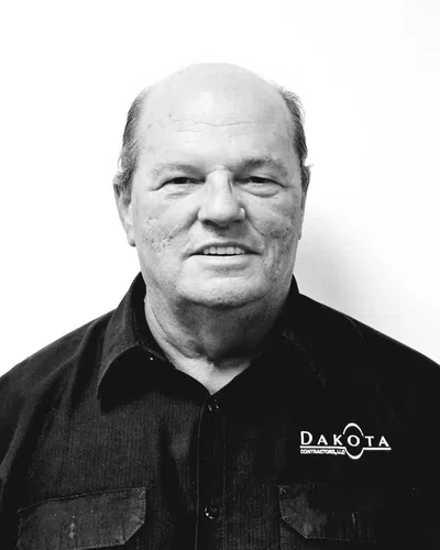 Older man with short hair wearing a black Dakota branded shirt standing against a white background.