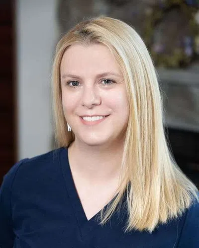 Smiling blonde woman in navy top with straight hair posing in a softly lit indoor setting