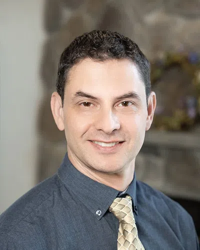 Smiling man with short dark hair wearing a blue shirt and beige patterned tie against blurred background