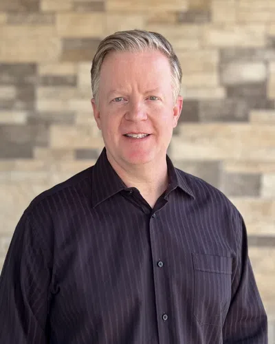 Middle-aged man with light hair wearing a dark striped button-up shirt standing in front of a stone wall.