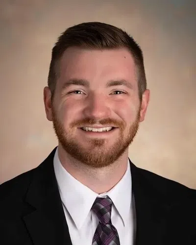 Smiling young man with short hair and beard wearing a black suit and patterned tie against a neutral background.