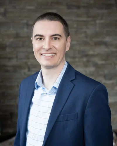 Smiling man in blue suit jacket and checkered shirt standing against a stone wall background.