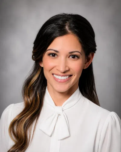 Professional woman with long dark hair and white blouse smiling against a gray backdrop.