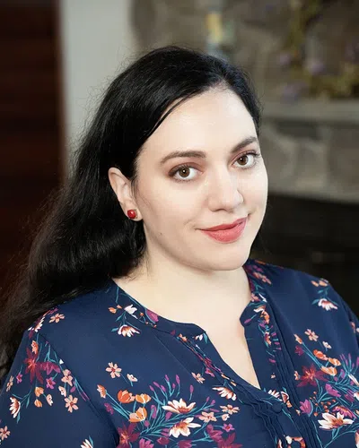 Smiling woman with dark hair wearing a navy floral blouse and red earrings in an indoor setting