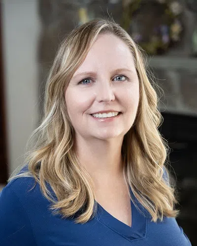 Smiling woman with wavy blonde hair wearing a blue top in a softly lit indoor setting.