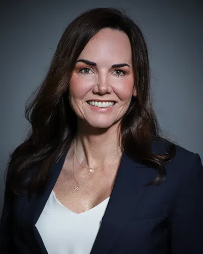 Smiling woman with long brown hair wearing a navy blazer and white top against a gray background.